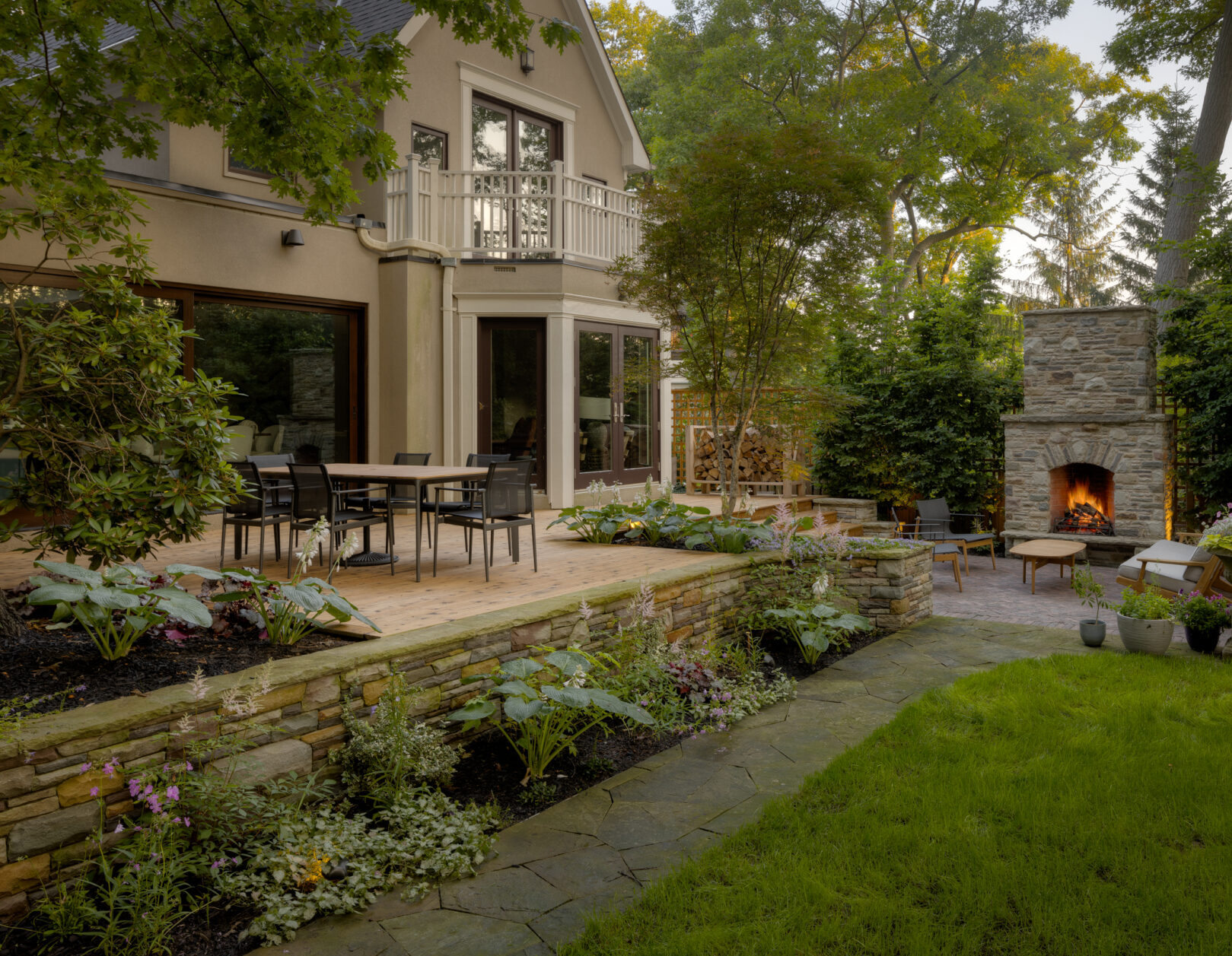 A backyard of a large house with a patio, and stone retaining wall and outdoor fireplace.